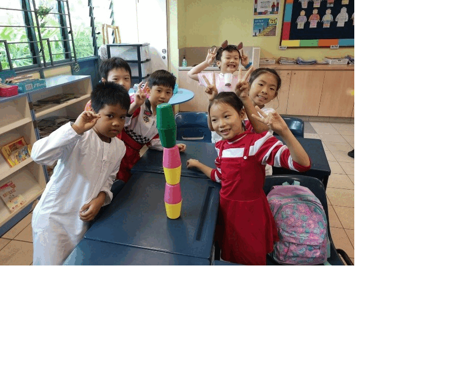 Five young children smile and pose with peace signs around a desk holding a stack of colorful cups in a classroom.